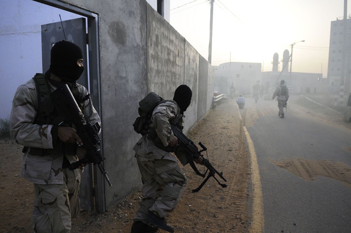 Two masked Israeli counter-terrorism officers in desert camouflage carry rifles along the wall of a mock village at dawn, a third walking ahead down the road.