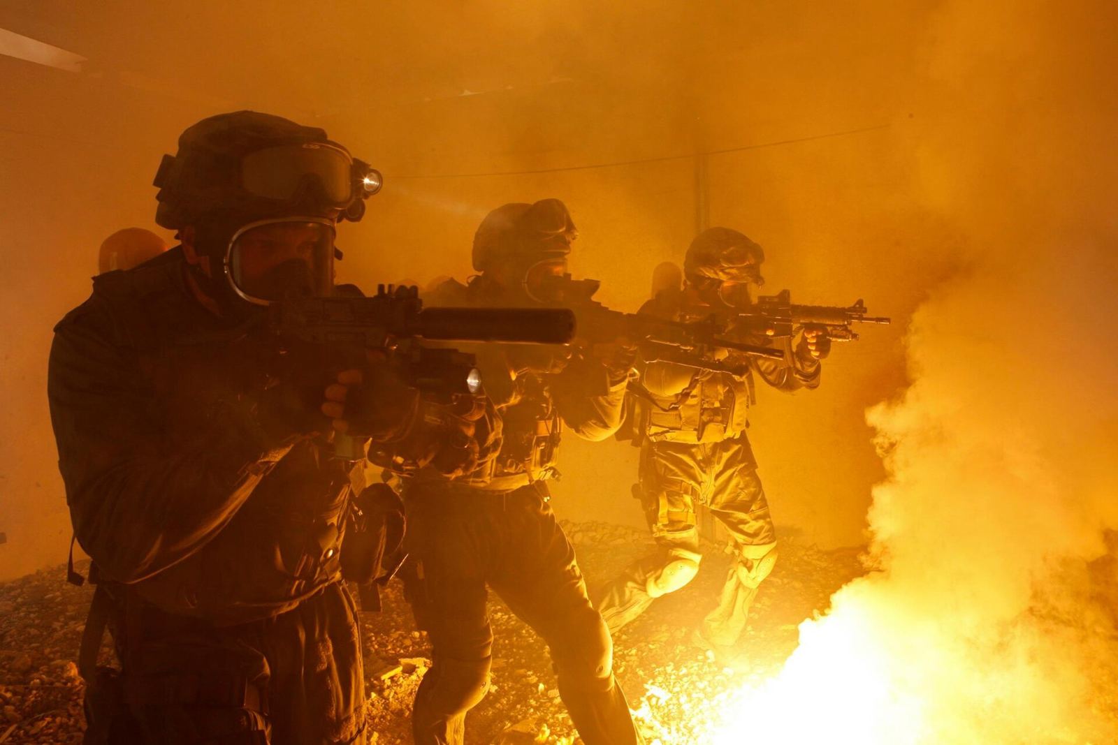 Three Israel Police counter-terrorism officers in gas masks and helmets advance with rifles raised through a smoke-filled room lit orange by fire.