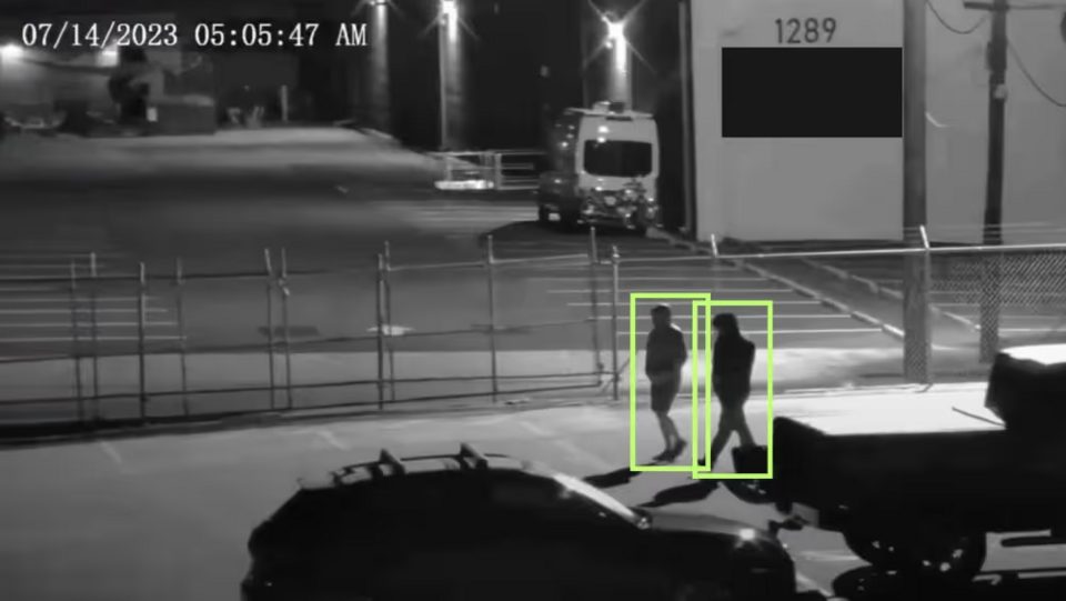 Black-and-white security camera frame, time-stamped 07/14/2023 05:05:47 AM, of two people walking along a fenced lot at night, each outlined by a green tracking box drawn by the software.