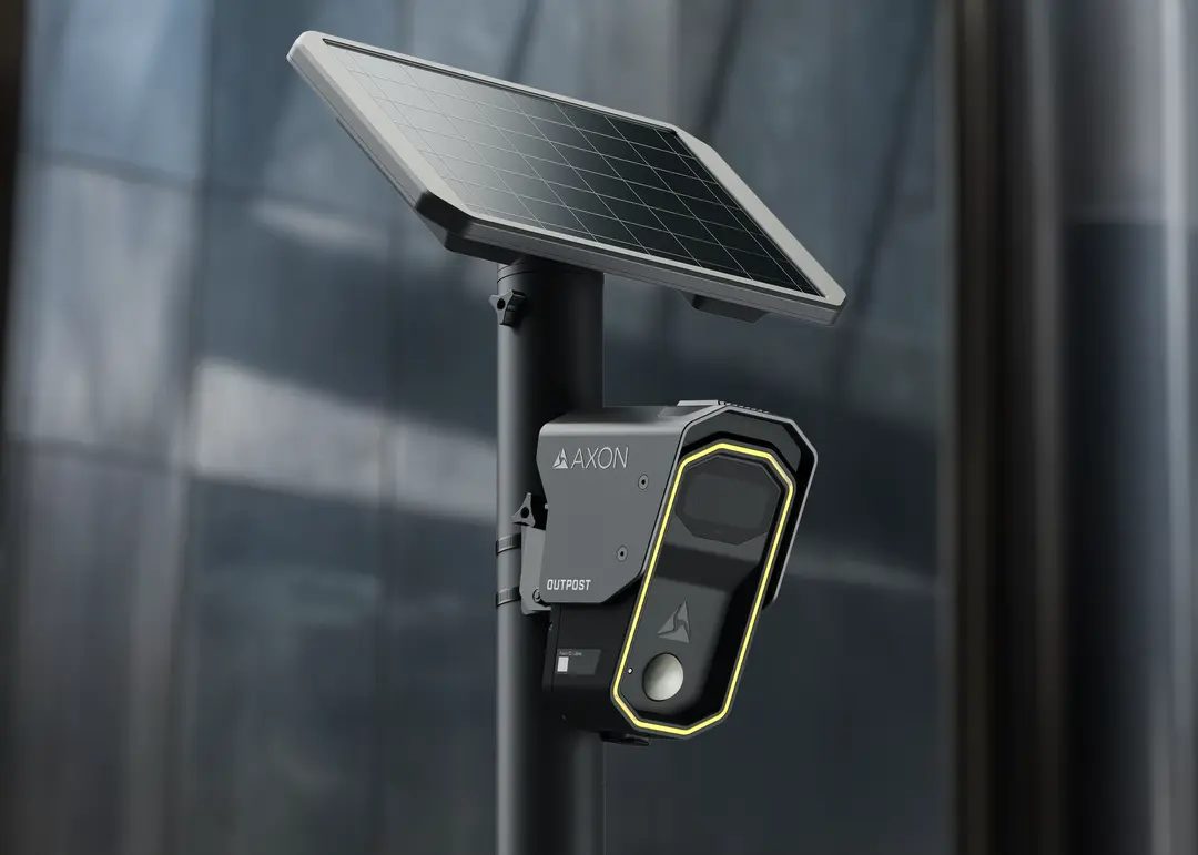 An Axon Outpost license-plate reader, a dark angular camera unit with a yellow-edged lens housing, mounted on a pole beneath a solar panel.