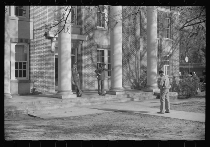 A 1941 black-and-white photograph: a man pauses on a sunlit courthouse walk while two soldiers stand at the columned entrance.